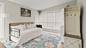 A bedroom with a bed, a window with blinds, a bedside lamp, a floral rug, and a wardrobe.