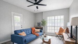 A living room with a blue sofa, a glass coffee table, a ceiling fan, and a large window with blinds.