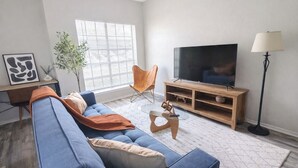 A living room with a blue sofa, a TV on a wooden stand, a floor lamp, and a window with blinds.
