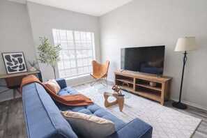 Ein Wohnzimmer mit einem blauen Sofa, einem Fernseher auf einem Holzschrank, einer Stehlampe und einem Fenster mit Jalousien.