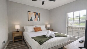 A bedroom with a bed, two bedside tables, a ceiling fan, a window with blinds, and a painting on the wall.