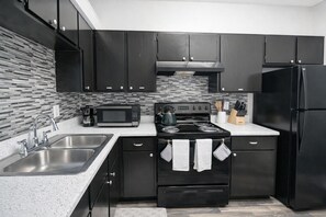 A modern kitchen with dark cabinets, a stainless steel sink, a microwave, a stove, and a refrigerator. - Chic 1BR Retreat | Gym, Pool & Free Parking (Irving)