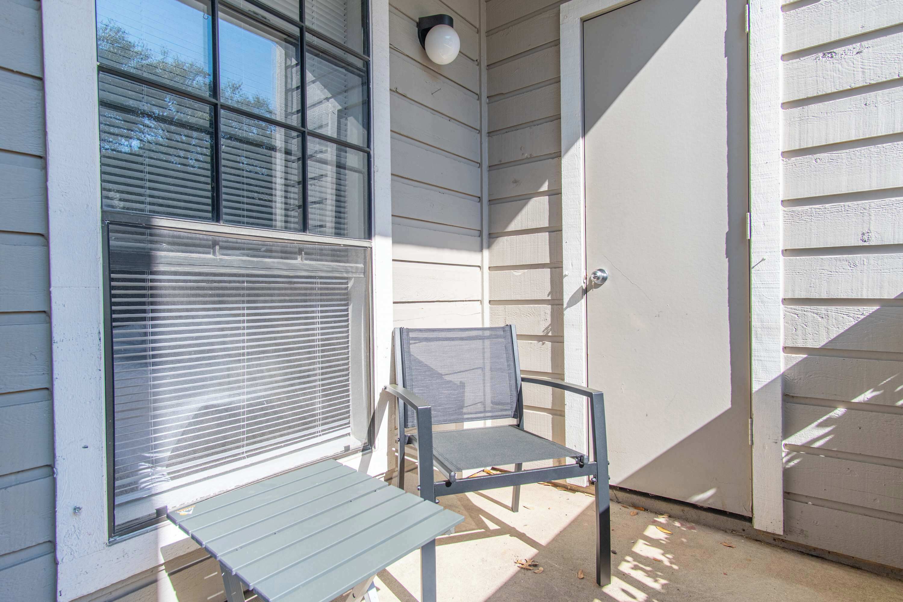 A small outdoor seating area with a metal table and chair, a window with blinds, and a door.