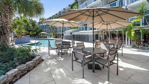 Un espace aménagé au bord de la piscine, avec des sièges et des parasols, une piscine, et un bâtiment doté de balcons.