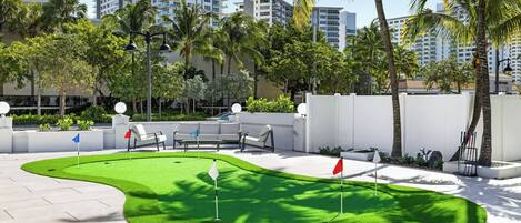 A golf putting green with white and red flags, surrounded by palm trees and modern buildings.
