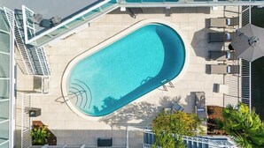 Una piscina en la azotea con barandilla de cristal, mobiliario exterior y palmeras.