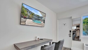 Ett modernt rum med en platt-TV som är monterad på väggen, ett skrivbord i trä, två stolar och en dörr som leder ut till en balkong med utsikt över poolen.