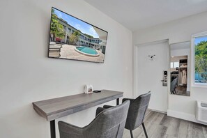 Ein modernes Zimmer mit Flachbildfernseher an der Wand, einem Holztisch, zwei Stühlen und einer Tür zu einem Balkon mit Blick auf den Pool.