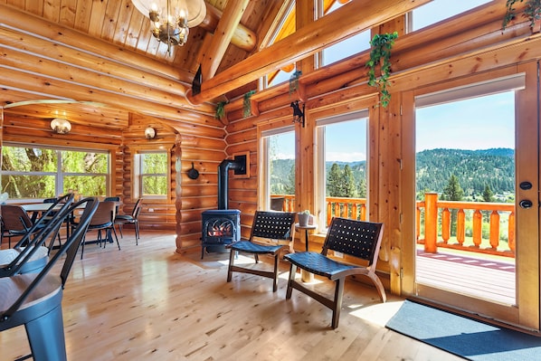 A log cabin interior with a fireplace, wooden furniture, large windows, and a balcony with a view of the mountains.