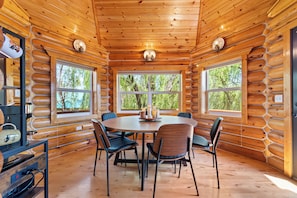 A log cabin interior with a round dining table, six chairs, and large windows offering a view of trees.