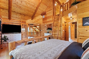 Ein Schlafzimmer in einer Blockhütte mit Bett, Schminktisch, Fernseher und einer Treppe zu einer oberen Ebene.
