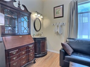 Une pièce comprenant une commode en bois foncé, un miroir rond, une coiffeuse avec un lavabo, un canapé en cuir noir et une fenêtre avec des rideaux.