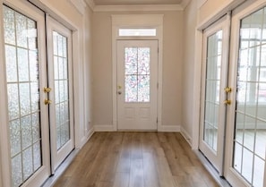 Un pasillo con piso de madera, una puerta blanca con un vitral y puertas de vidrio con diseño de cuadrícula.