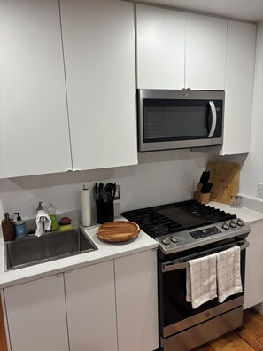 Moderni keittiö, jossa on valkoiset kaapit, ruostumattomasta teräksestä valmistettu liesi, mikroaaltouuni ja puinen leikkuulauta.