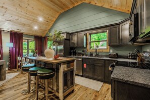 Une cuisine avec un plafond en bois, des meubles sombres, un îlot central, des tabourets de bar et une vue sur la verdure à l’extérieur.