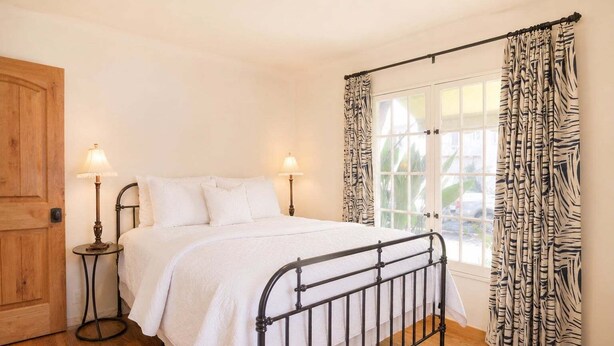 Une chambre à coucher avec un lit, deux lampes de chevet, une porte en bois, une fenêtre avec des rideaux et un plancher en bois.