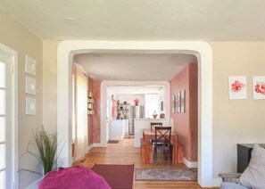 Ein Wohnzimmer mit Sofa, Couchtisch und Fernseher. Es gibt einen Essbereich mit Tisch und Stühlen. Der Raum hat einen Holzboden und ein großes Fenster.