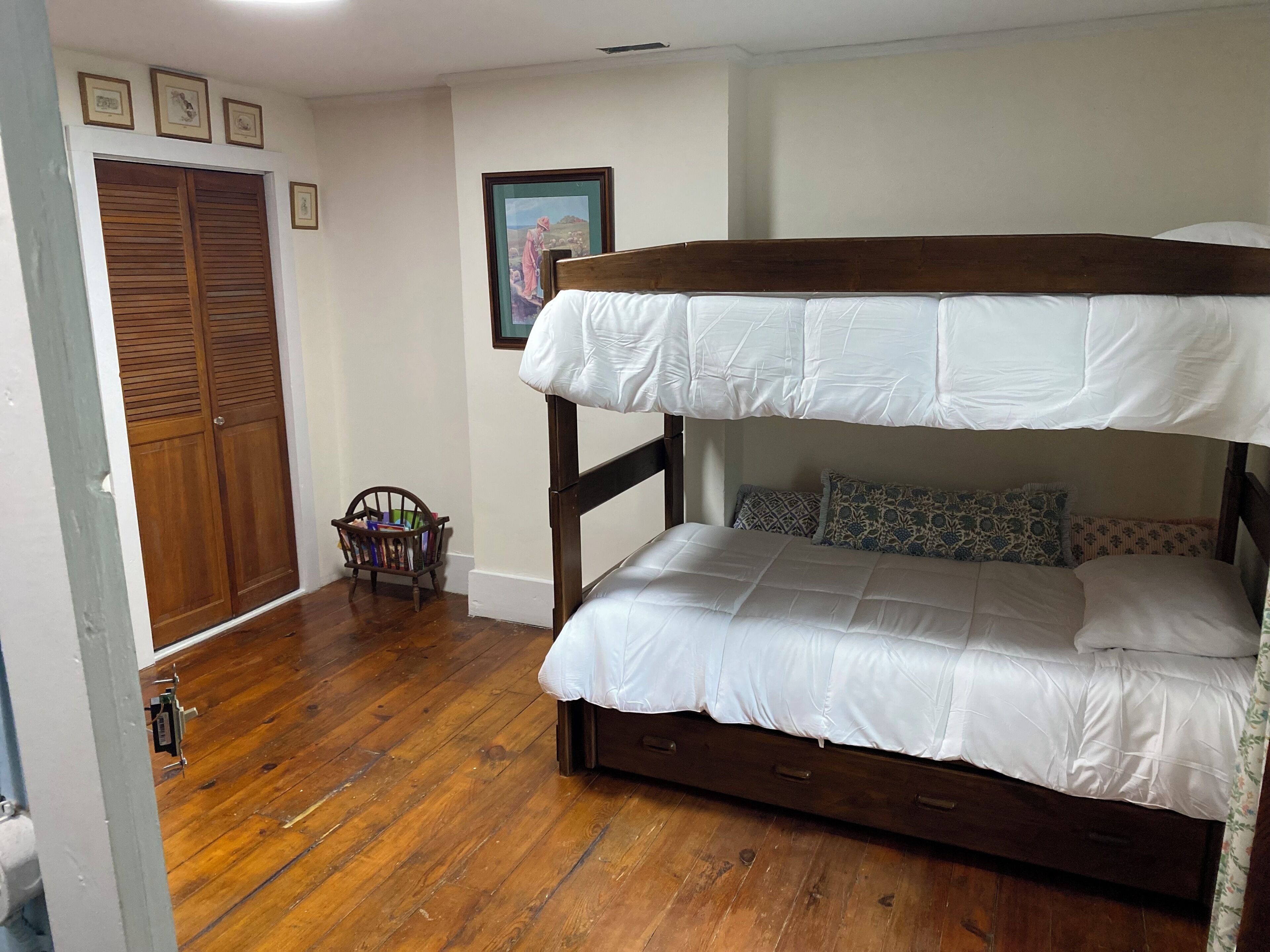 Una cama litera con estructura de madera y ropa de cama blanca, una puerta de madera con un pequeño ventanal y una pared decorada con marcos de fotos.