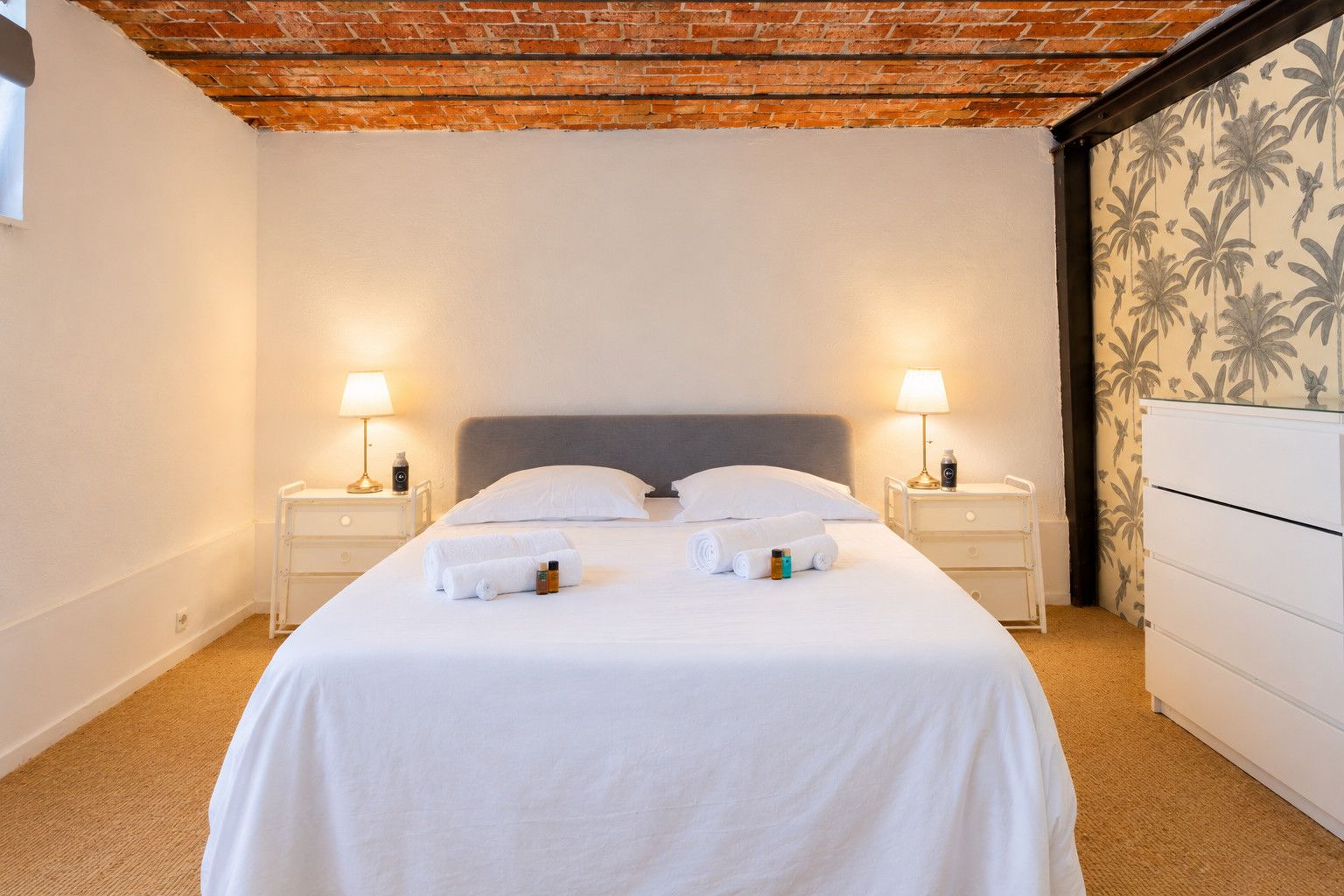 A hotel room with a bed, two bedside tables with lamps, a white dresser, and a brick ceiling.