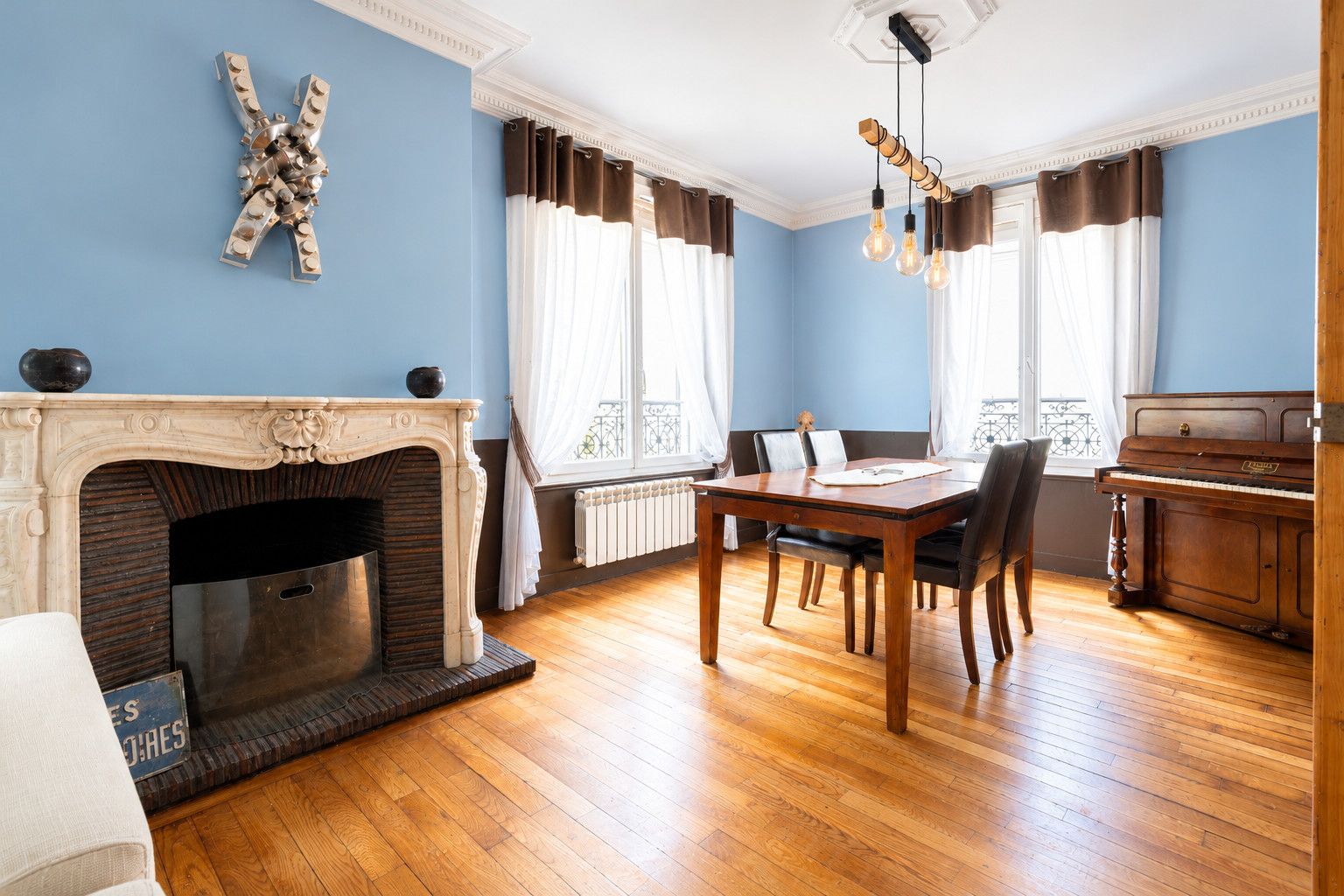 Une pièce avec une cheminée, une table à manger, des chaises, un piano et une grande fenêtre avec des rideaux.
