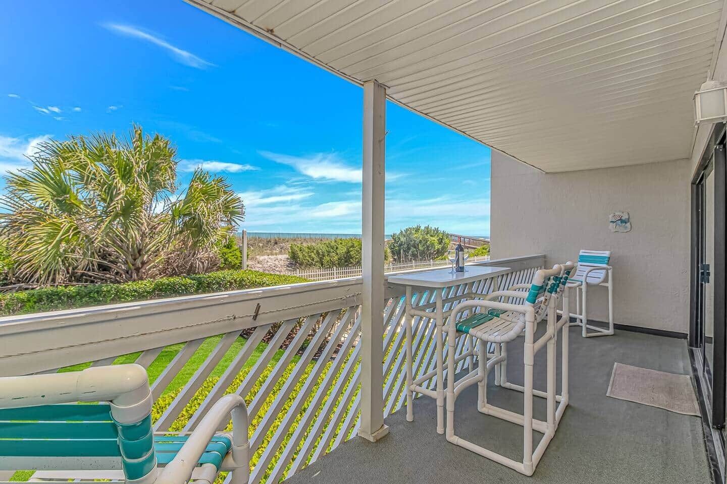 A balcony with a view of the beach, a table with chairs, and a small table with a bottle.