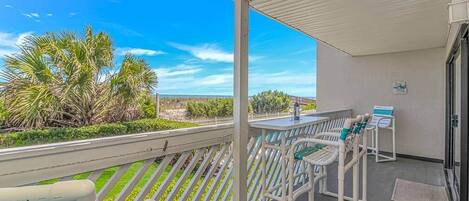 A balcony with a view of the beach, a table with chairs, and a small table with a bottle.