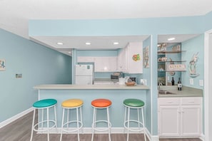 A kitchen with a central island, white cabinets, and colorful stools. - 3BR Oceanfront Condo | Pool | Main Street (North Myrtle Beach)