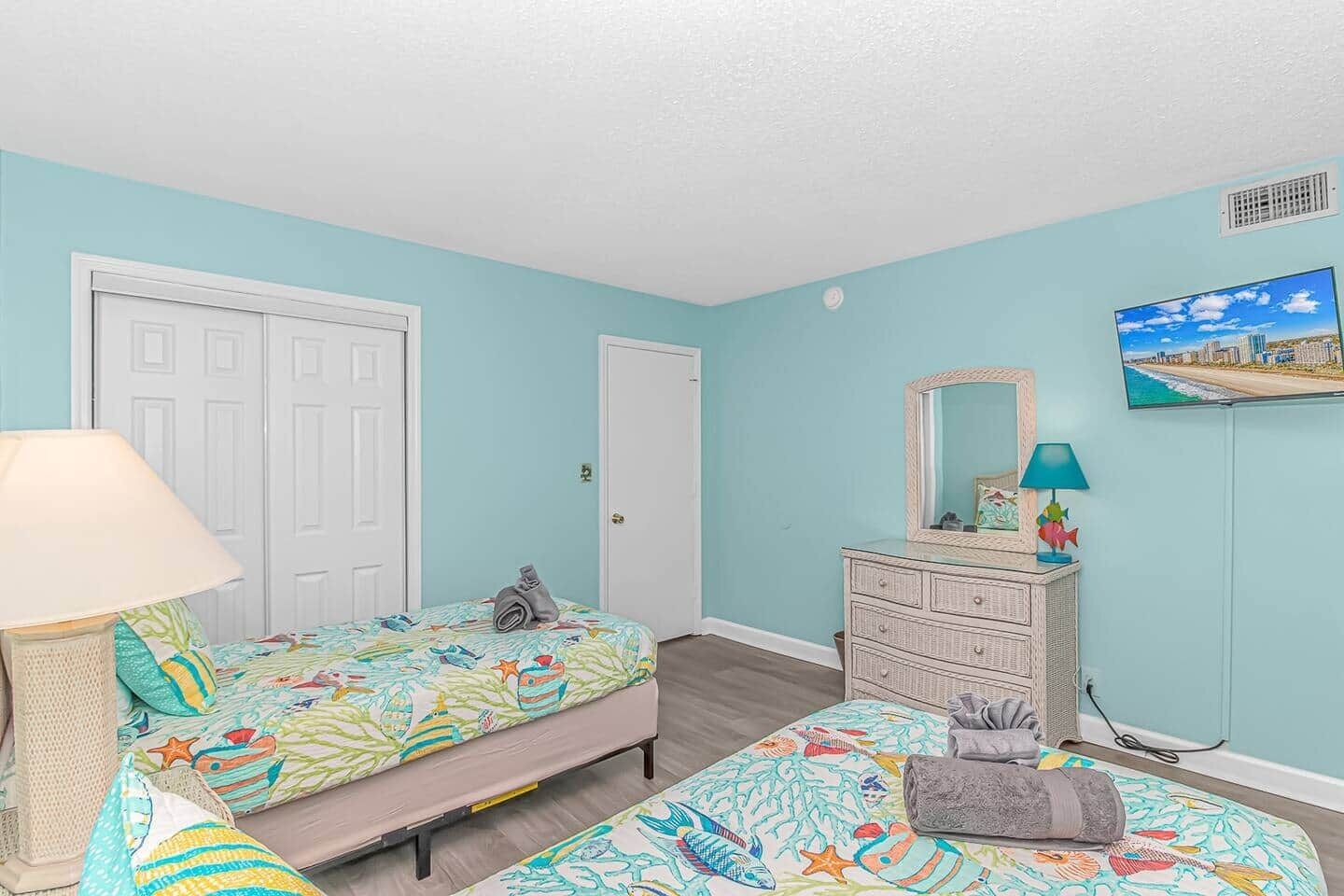 Une chambre avec deux lits, une commode, une lampe et un téléviseur fixé au mur.