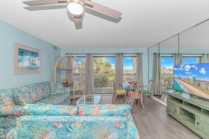 A living room with a sofa, a dining table with chairs, a TV, and a ceiling fan. - 3BR Oceanfront Condo | Pool | Main Street (North Myrtle Beach)