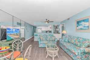 A living room with a sofa, chairs, a TV, and a kitchen area. - 3BR Oceanfront Condo | Pool | Main Street (North Myrtle Beach)