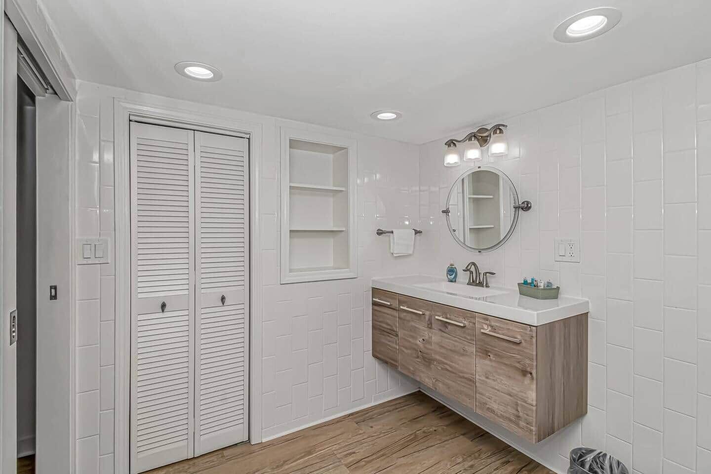 A bathroom with a white tiled wall, wooden vanity, a mirror with lights, and a wooden floor.