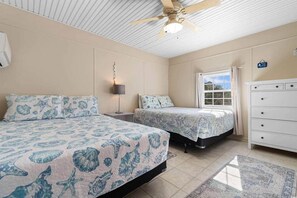 Une chambre avec deux lits, un ventilateur de plafond, une table de chevet avec une lampe, une commode et une fenêtre donnant sur un beau paysage.