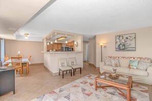 A living room with a sofa, coffee table, dining area with chairs, and a kitchen area with cabinets and appliances. - Updated 3BR w/ Oceanviews on MB Boardwalk! (Myrtle Beach)