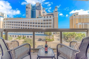 A balcony with wicker chairs and a table, overlooking a cityscape with modern buildings and a parking lot. - Updated 3BR w/ Oceanviews on MB Boardwalk! (Myrtle Beach)