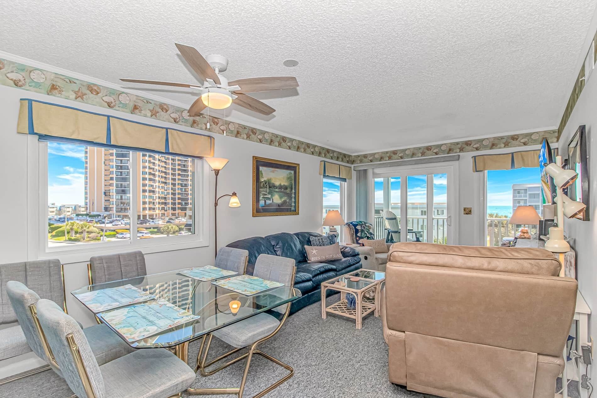 A living room with a glass coffee table, a sofa, a chair, a ceiling fan, and large windows with a view of buildings.