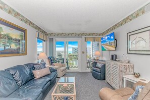 Ein Wohnzimmer mit einer Ledersofa, einem Glastisch, einem beige Armlehnstuhl, einem Fernseher und einem Balkon mit Blick auf die Stadt.