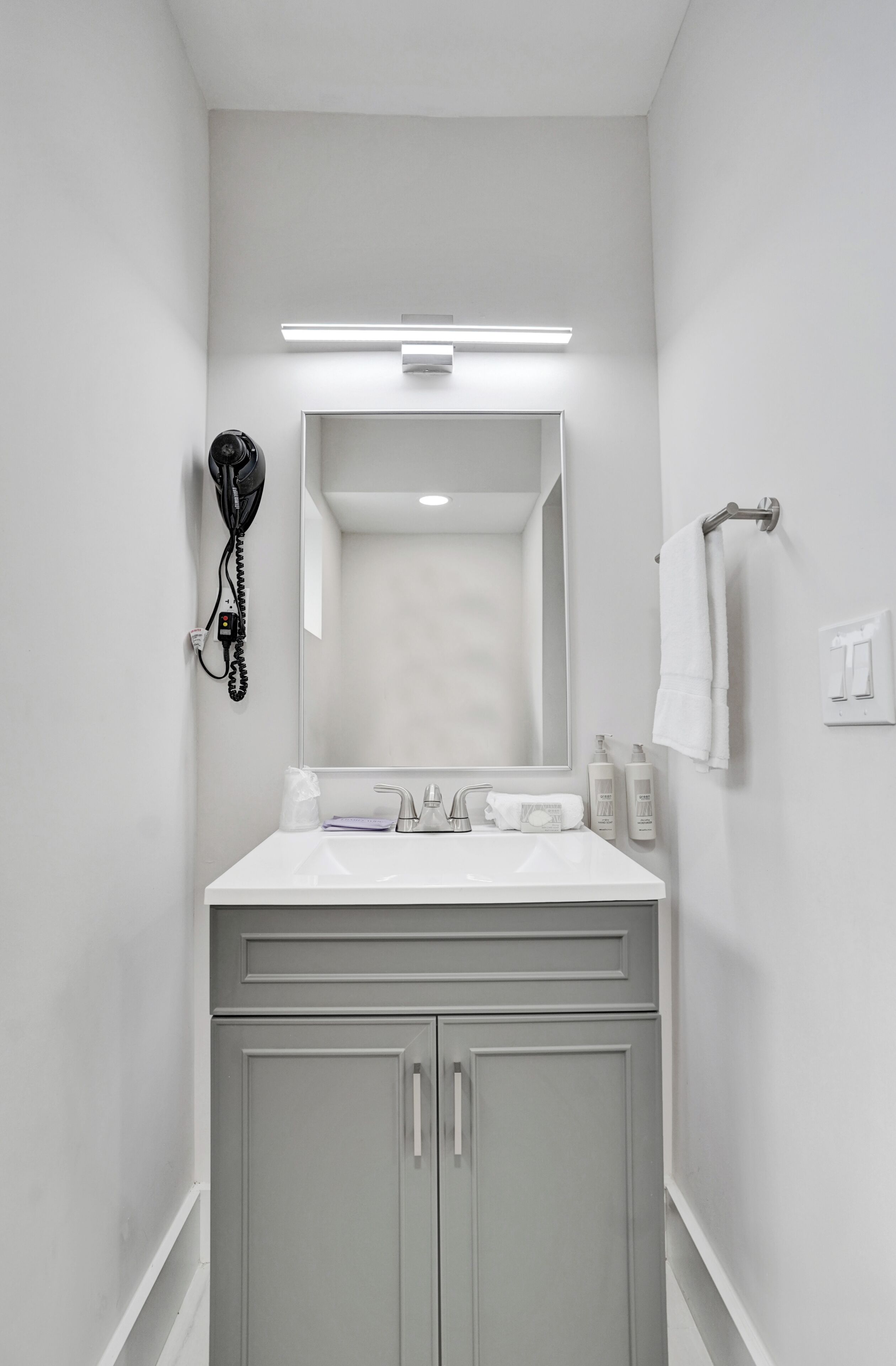 A bathroom with a white sink, mirror, and cabinets. There is a hairdryer on the wall and a towel rack.