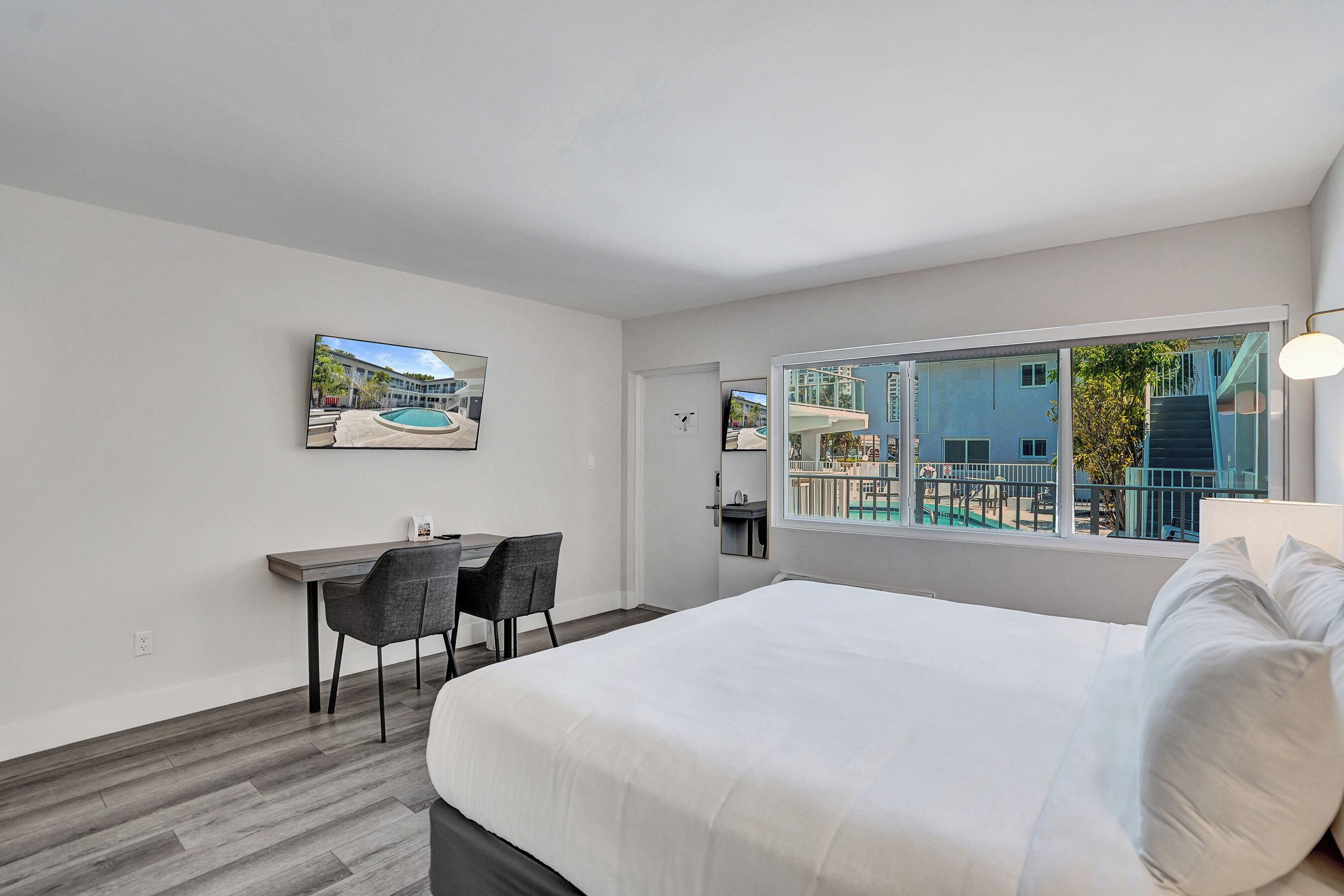 Habitación de hotel con cama, escritorio, sillas y un amplio ventanal con vistas a una piscina y edificios.