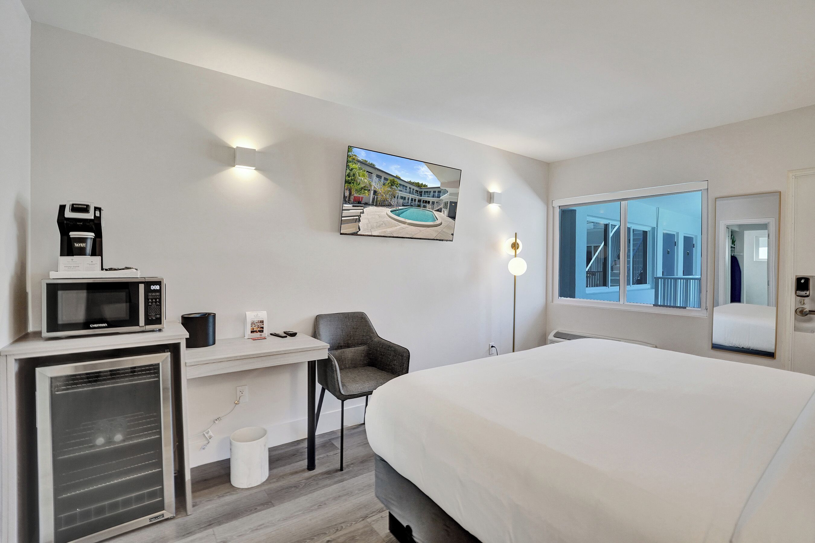 Habitación de hotel con cama, escritorio con microondas, una silla, televisor montado en la pared y ventana con vista a un edificio.