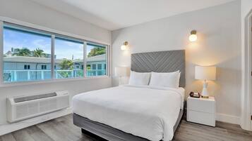 Un dormitorio moderno con una cama grande, dos lámparas de noche, una ventana con vista a edificios y un aire acondicionado integrado.