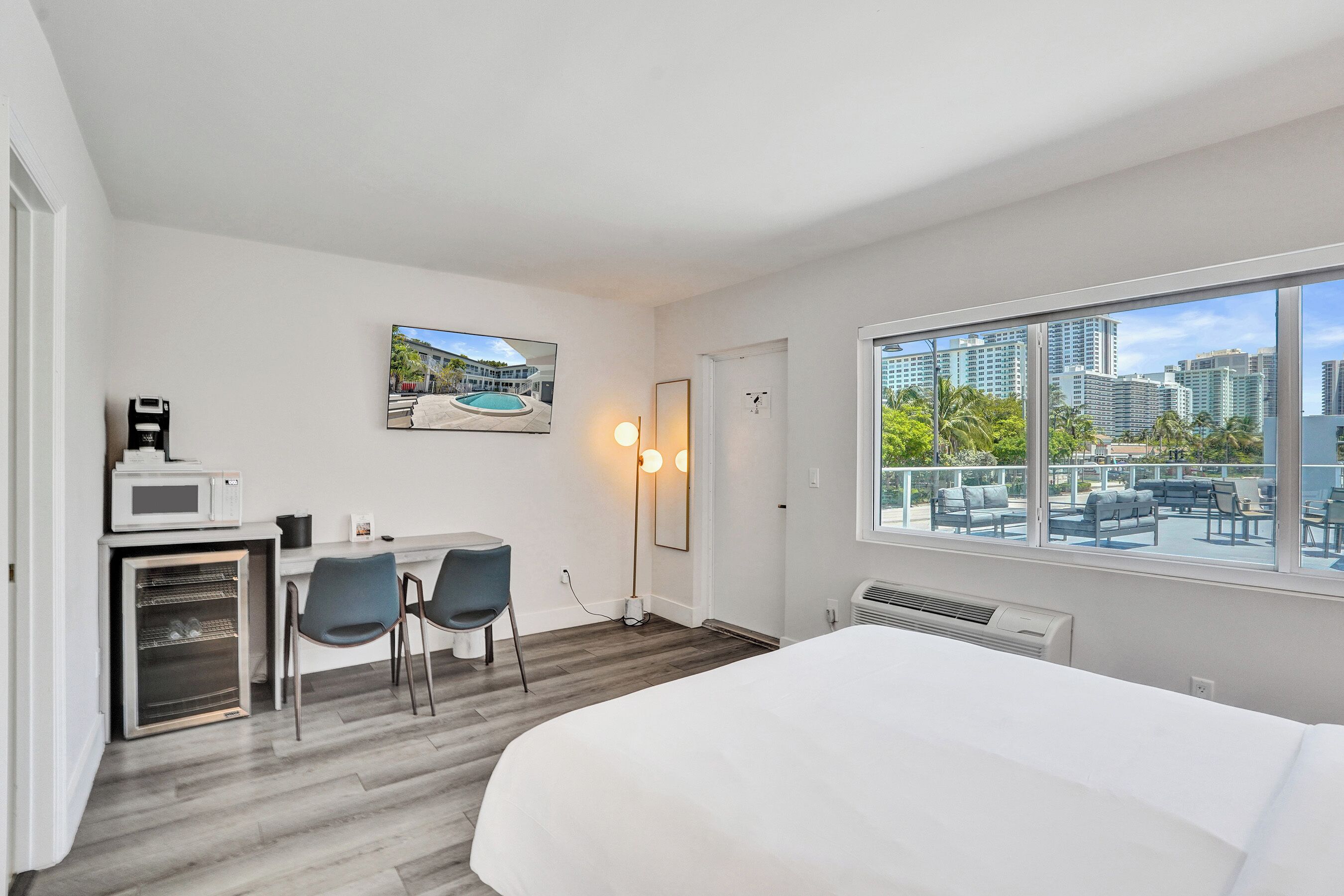 A hotel room with a bed, a desk with a microwave, two chairs, a large window with a city view, and a framed picture on the wall.
