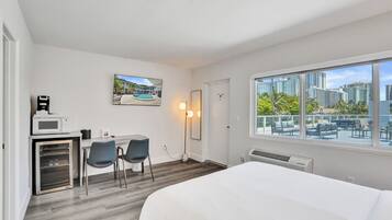 A hotel room with a bed, a desk with a microwave, two chairs, a large window with a city view, and a framed picture on the wall.