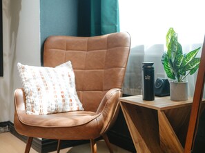 Una silla de cuero marrón con un cojín estampado, un vaso negro y una planta en maceta sobre una mesa de madera.
