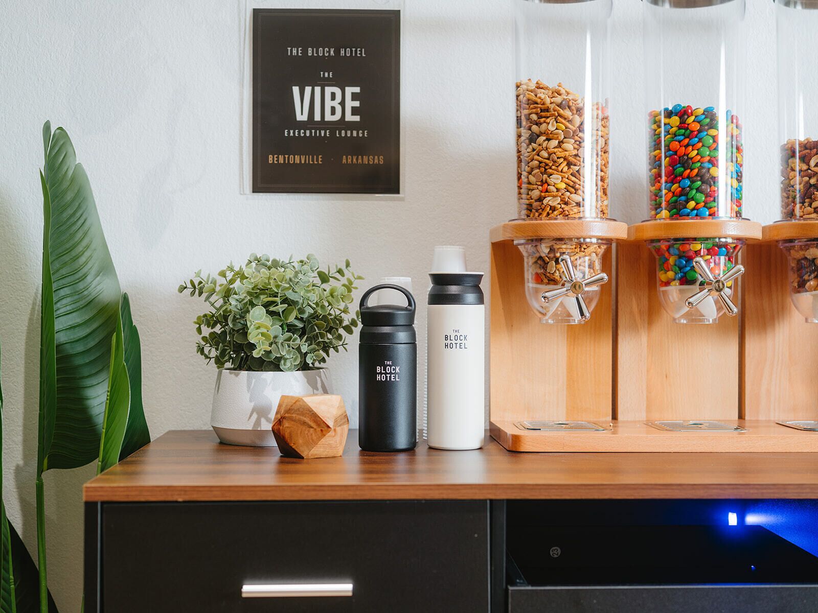 A hotel bar area with a sign reading 'The Block Hotel The Vibe Executive Lounge Bentonville Arkansas', a potted plant, a water bottle, and jars of snacks.