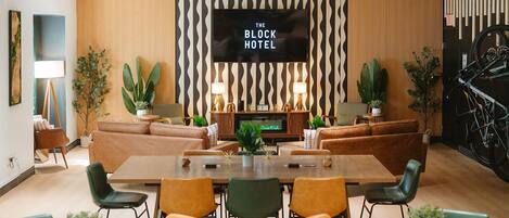 Un vestíbulo de hotel con una gran pantalla de televisión mostrando 'THE BLOCK HOTEL', un área de descanso con sofás y sillas, y una mesa de comedor con sillas.