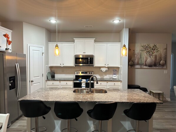 A modern kitchen with white cabinets, a stainless steel refrigerator, a built-in oven, a microwave, a sink, and bar stools.