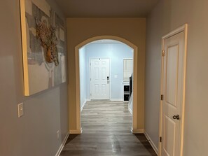 A hallway with a white door at the end, a light switch on the wall, and a partial view of a room with a window and blinds. - Spacious 4BR Family Retreat Near Whitewater & Airport, pets allow (Charlotte)