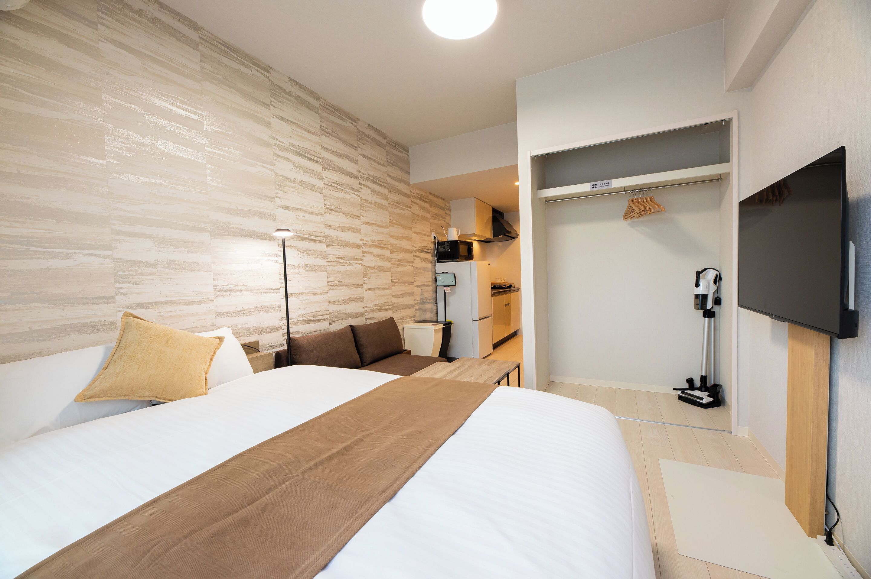 A modern hotel room with a large bed, a wardrobe, a television, and a kitchenette.