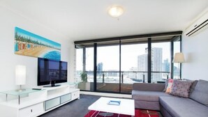A modern living room with a large window, a flat-screen TV, a sofa, and a glass coffee table. - Exclusive Stays - Yarra Condos (Southbank)