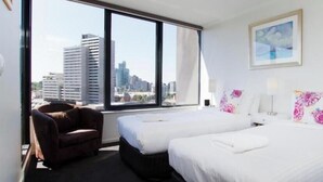 A hotel room with two beds, a chair, a large window with a city view, and a framed picture on the wall. - Exclusive Stays - Yarra Condos (Southbank)
